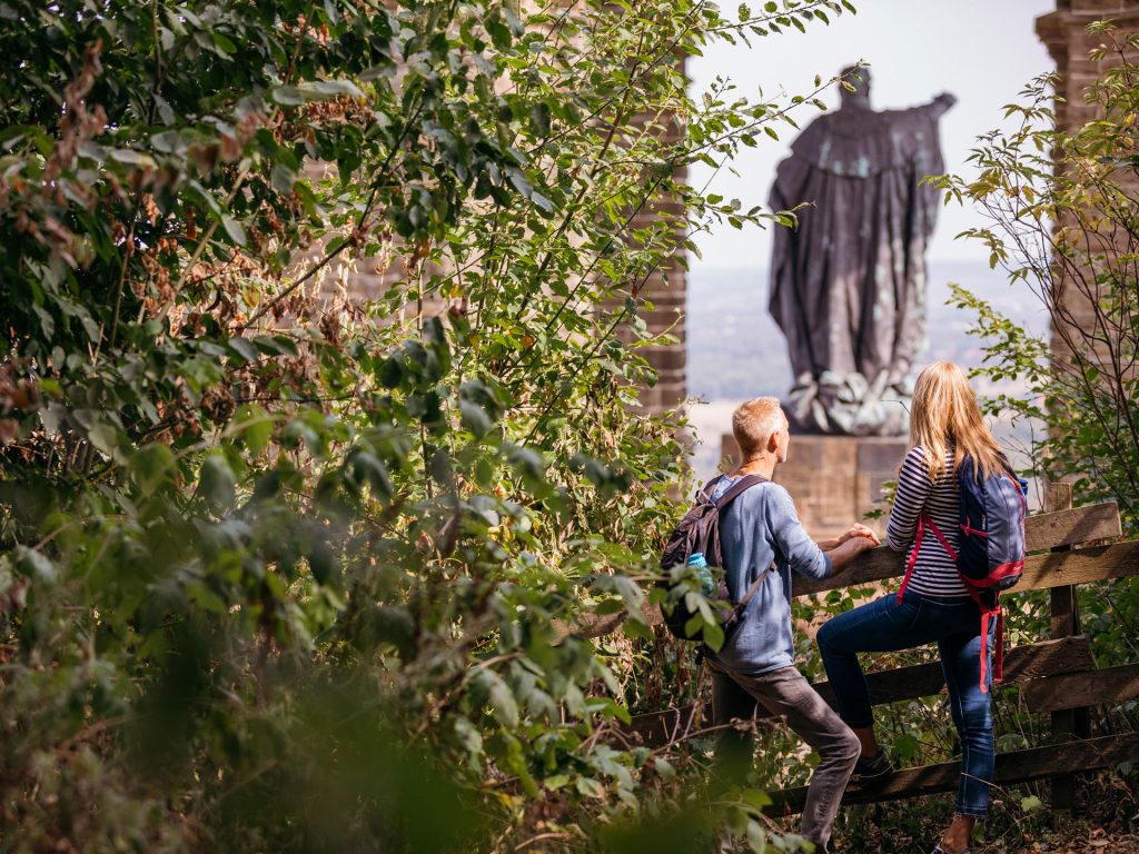 Zwei Wandernde mit Rucksack steigen eine Bank am Waldrand hinauf und blicken auf das Kaiser-Wilhelm-Denkmal in Porta Westfalica. Die Szene steht für Ausflüge mit Bus und Bahn in OWL.