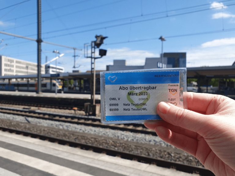 Hand hält ein übertragbares OWL-Abo-Ticket am Bahnsteig eines Bahnhofs – unterwegs mit dem WestfalenTarif in der Region TeutoOWL.