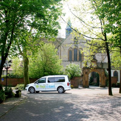 flex-taxi-Fahrzeug steht auf einem gepflasterten Platz vor dem historischen Kloster Marienfeld in Harsewinkel – flexible Mobilität vor Ort.